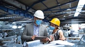 Two employees collaborate on capital equipment planning in a healthcare factory, surrounded by machinery and resources.