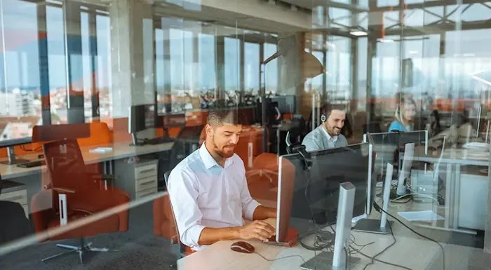 Three co-workers share office space, working on computers at a hot desk setup.
