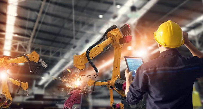 An engineer operating a tablet while overseeing robotic arms welding in a factory